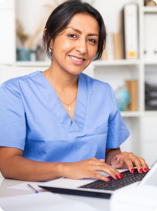 A woman in blue scrubs is typing on a laptop.